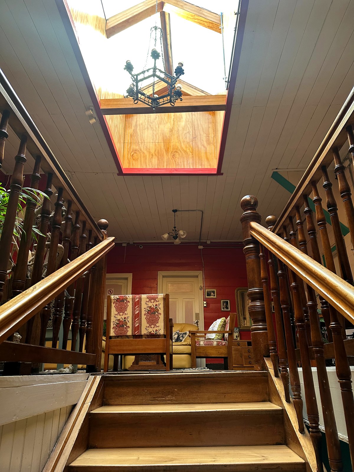 Escalera de madera original de la casa, con claraboya, candelabro de hierro y salón en tonos rojos al fondo